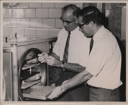 A black and white photograph of B. W. Calnek and S. B. Hitchner examining samples of the Marek's Disease vaccine in a lab in 1970.