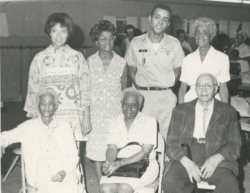 A group of family memebers next to eachther, one of which if wearing a military unfiform, and three of which are sitting in chair in front of the rest standing.