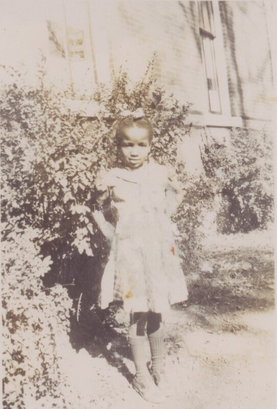 Young child in the yard in front of a bush poses for a photo.