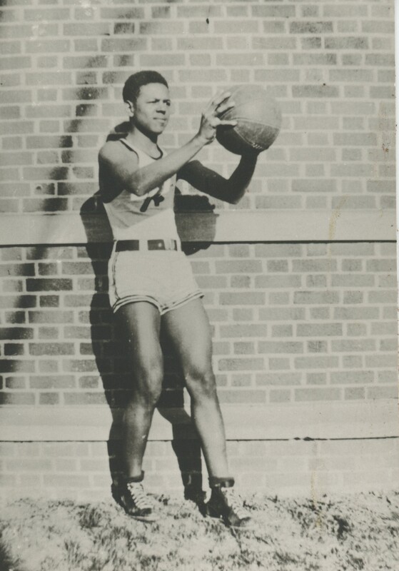 A basketball player wearing their basketball uniform holding a basketball away from their body stnding in front of a brick wall.