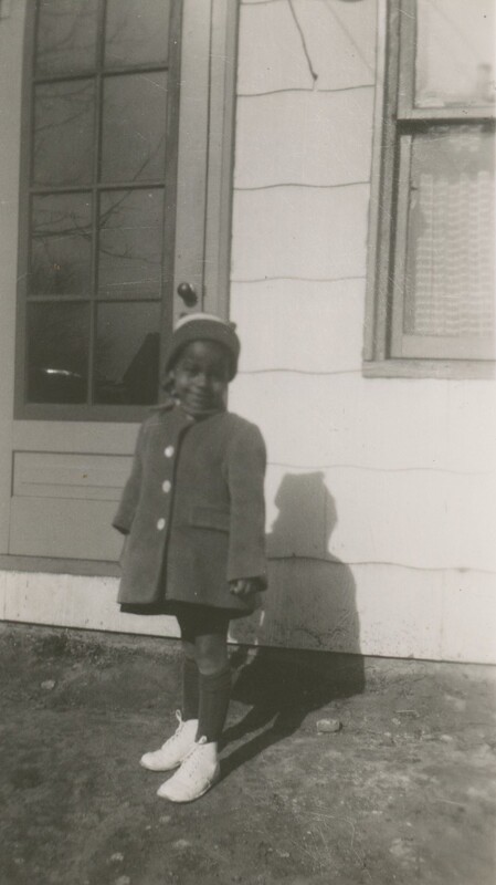 A little child stood by a front door of a house wearing a coat dress and a hat smiling.