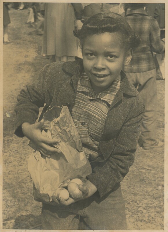 A young child holding some eggs and a brown paper bag with some people stood in the backgrounds.