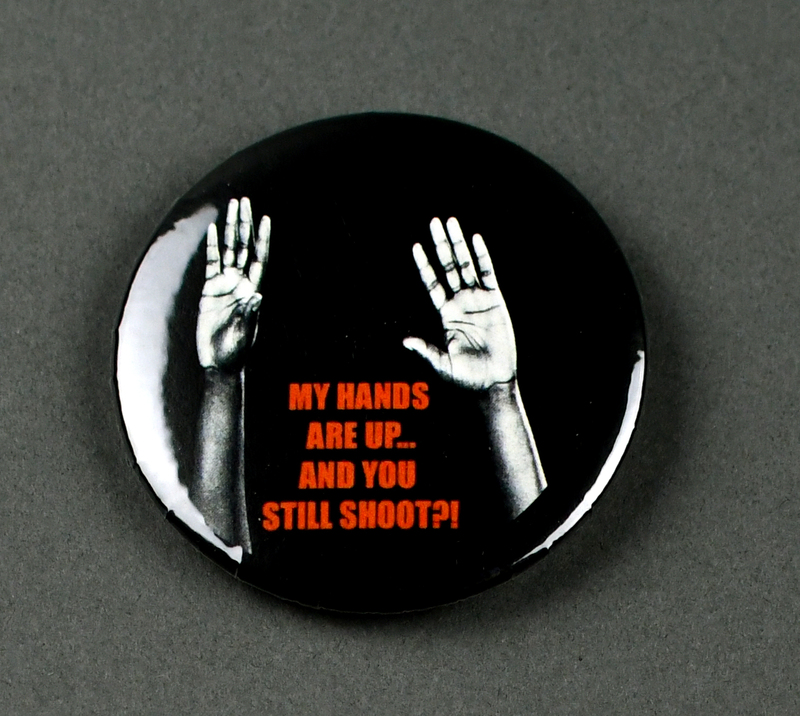 Button with safety pin clasp at posterior. Graphic at anterior printed on paper and crimped under transparent plastic covering. "MY HANDS ARE UP... AND YOU STILL SHOOT?!" in red on black background. Black and white image of two raised hands behind text.  

Button made and sold/distributed by Amani Community Services during the 2020 George Floyd Protests in Waterloo. Amani community services is a domestic violence and sexual assault prevention agency serving African Americans in Black Hawk and Linn Counties with offices in Waterloo and Cedar Rapids.