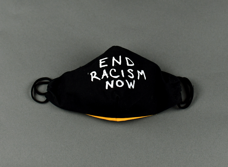 Covid mask of black fabric with orange fabric backing. Hand-made. Machine-stitched. “END RACISM NOW” in white acrylic paint at center anterior.   

Mask made by Maggie Dvorak of Cedar Falls. Worn by donor at the June 6 vigil in Overman Park, Cedar Falls, and the march from Clay Street to Overman Park, June 20, Cedar Falls.  Waterloo Courier photos show donor in mask at the June 6 vigil.

From donor Maggie Dvorak:

“I chose to contribute masks for several events in the Cedar Valley. These were events to bring community leaders and members together to show support for Black lives mattering, and to denounce systemic racism in our communities, and world wide.

Due to Covid 19, and my belief in the importance of doing our best to protect each other during a world wide pandemic, it was important to provide as many masks as necessary for participants to have available for use.

I hope to continue to donate and contribute my voice and my mask making abilities in any opportunity I am able. I will do this in hopes of supporting community health, and to support bringing an end to the systemic racism present in our world.”