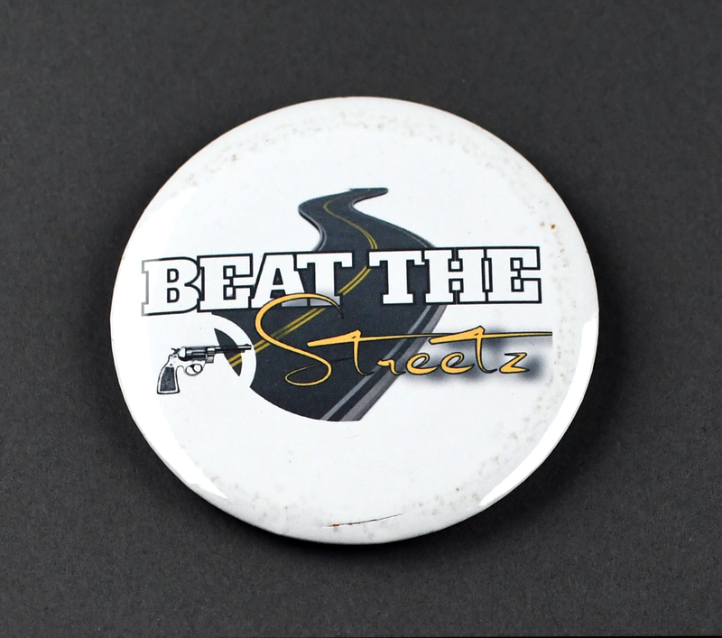 Button with safety pin clasp at posterior. Graphic at anterior printed on paper and crimped under transparent plastic covering. "BEAT THE Streetz" in white and yellow letters over a crossed-out revolver next to color representation of a street on a white background.

Beat the Streetz Foundation is a Waterloo nonprofit dedicated to providing education and resources to area youth, young adults, and adults for the prevention of gun violence. It’s founder, Raynelle Walker, is sister of local citizen Greg Walker, murdered 5/19/2018. The case remains unsolved. His nickname, “Streetz,” is the origin of the Foundation’s name.  
Beat the Streetz contributed to the organization of several Waterloo protests in 2020.