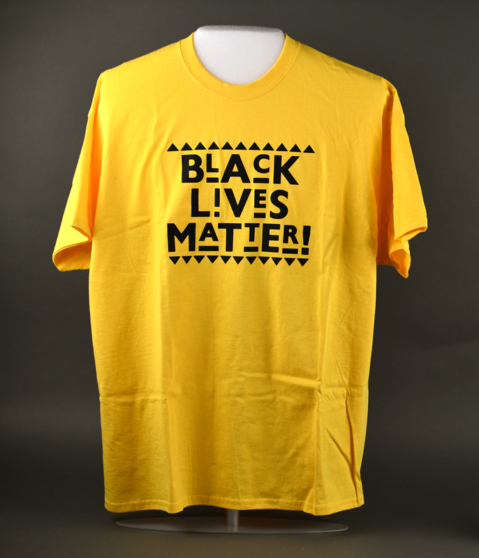 Crew-necked short-sleeve t-shirt of yellow cotton. Machine-stitched. “BLACK LIVES MATTER!” printed in black at center anterior. 
  
Printed and sold by Waterloo rapper Too Klean during the 2020 George Floyd Protests.