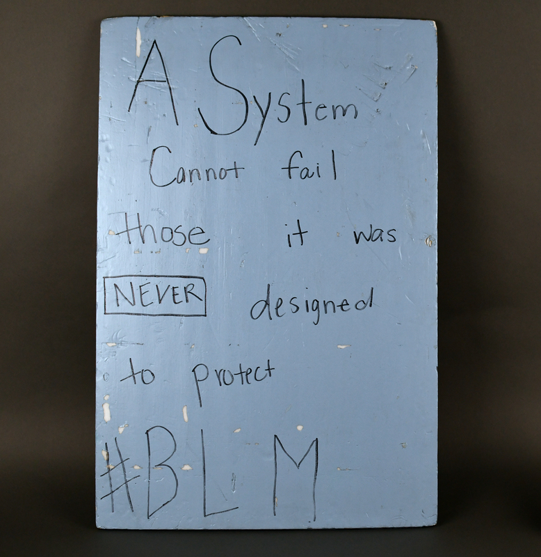 Black foamcore sign. Anterior and edges covered in blue acrylic paint. "A System Cannot fail those it was NEVER designed to protect, #BLM” handwritten in black felt-tip marker at anterior.     

In 2020, Nature Card was psychology major at the University of Northern Iowa in their junior year.  Card carried this sign in five protests in 2020:

- May 29, 2020, Veterans Memorial Hall to Lincoln Park, Waterloo.
- June 4, 2020, Lincoln Park, Waterloo.
- June 4, 2020, Police Station, Cedar Falls.     
- June 5, 2020, Evansdale.  
- August 31, 2020, Hawkeye Community College, Waterloo.