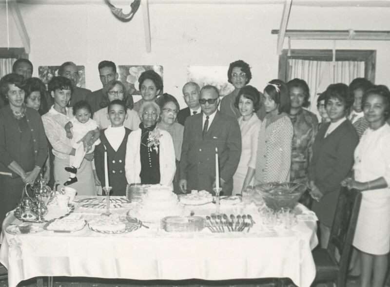 Multiple people are crowded around a table with a birthday cake and all of the utinsles for dining taking a photo at a party.
