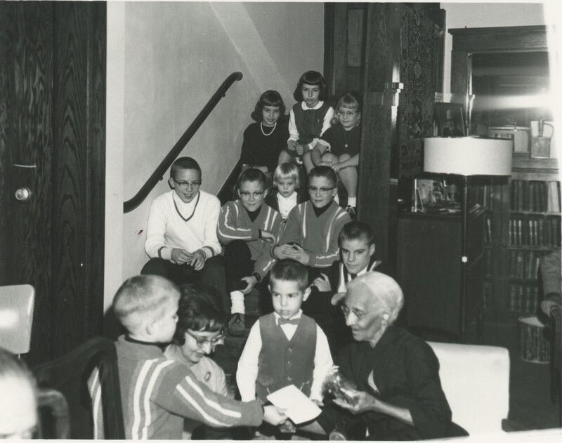 The birthday person is accepting gifts from some of the children while some of the others children watch from the staircase next to them.