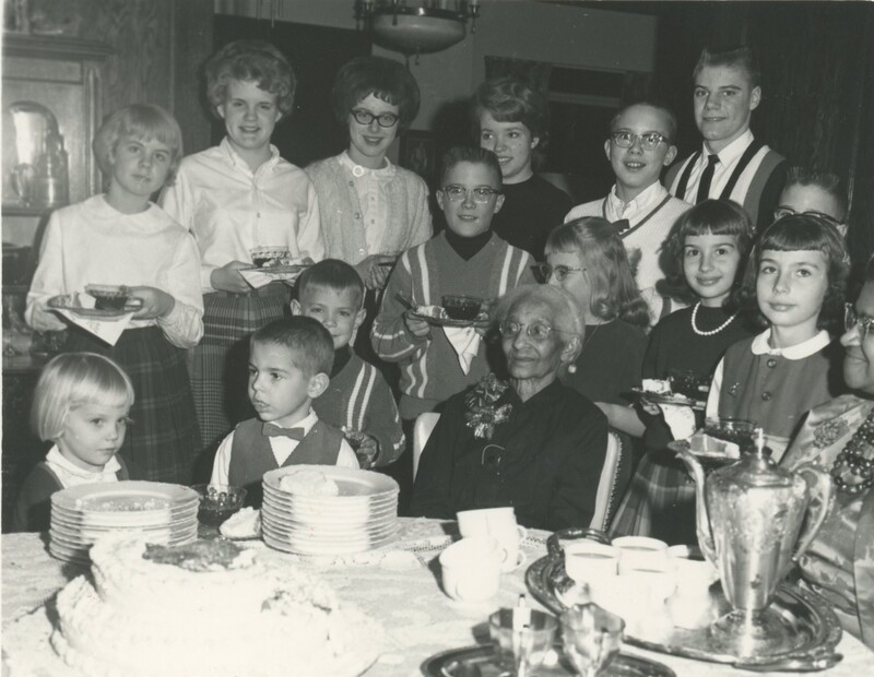 The children at the party ar stood around the birthday person eating cake and smiling for a photo.