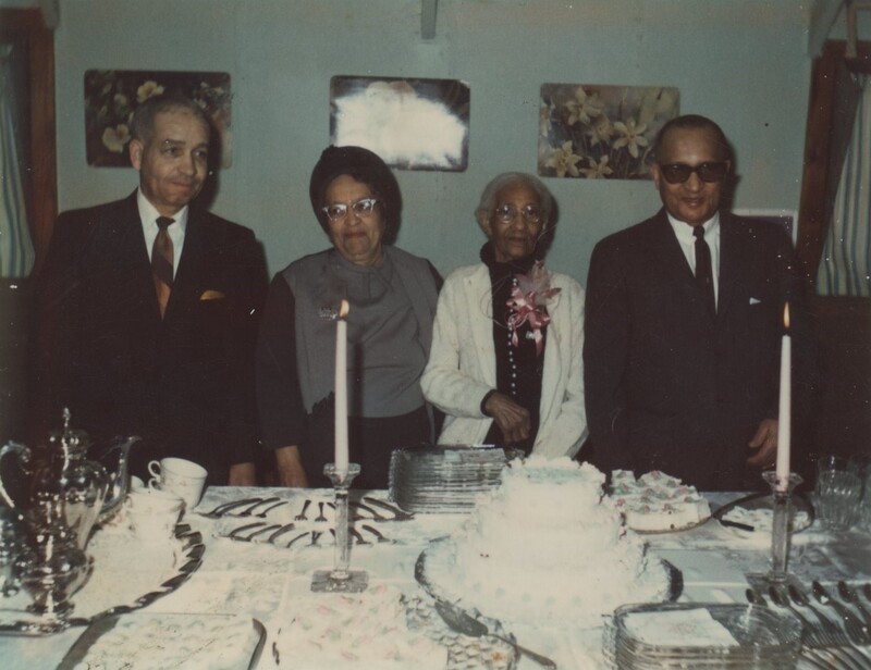 Four people stood behind a table with a birthday cake, cutlury, plates, and other utensils for dining.