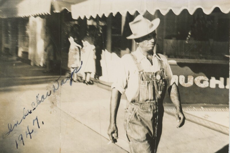 A person walking down the street in front of store fronts wearing a hat and overalls.