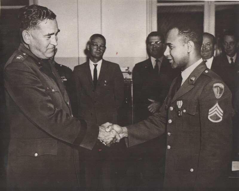 Two people in military uniforms shaking hands while four other people stand in the behind them.
