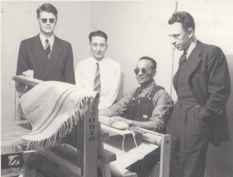 A person wearing dark glasses sat behand a loom machine with three people stood around for a photo.
