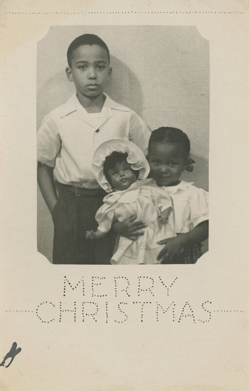 Two young children are stood next to eachother while one o them is holding a doll with the words "Merry Christmas" written at the bottom.