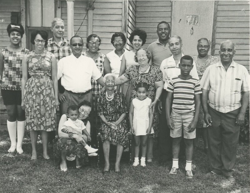 A family with multiple generations present are standing clumped together in the yard of a house right next to the house.