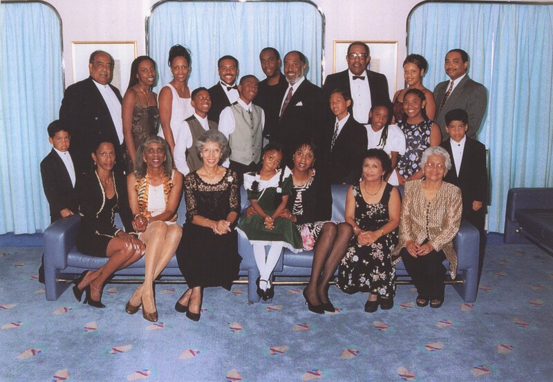 A large family are clumped together for a family photo while some of them are sat down on a sofa and the rest of stood behind the sofa.