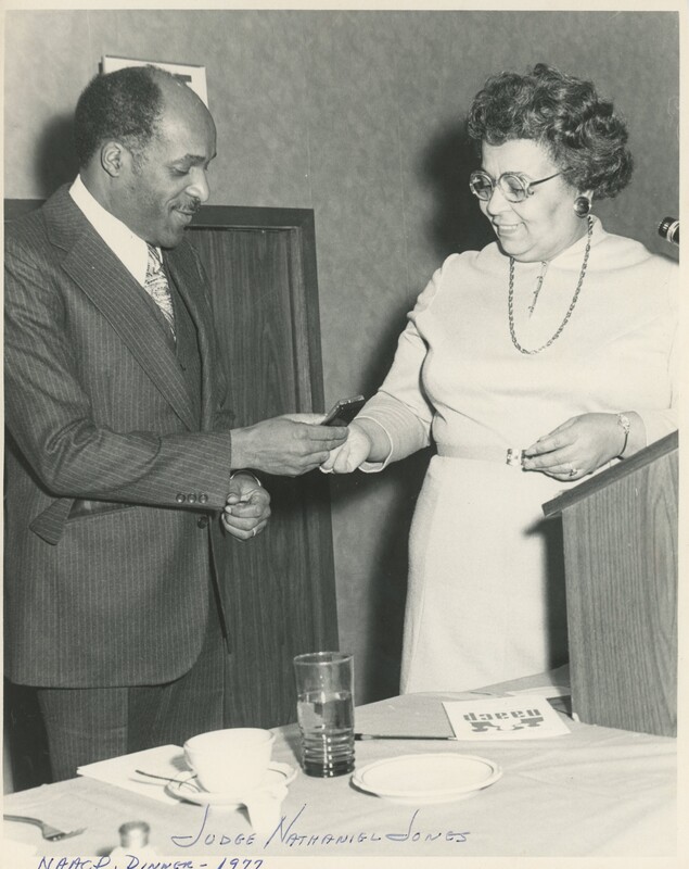 Two people stood across from each other while one is handing the other something stood behind a podium and a table with a glass of water and some cultery on it.