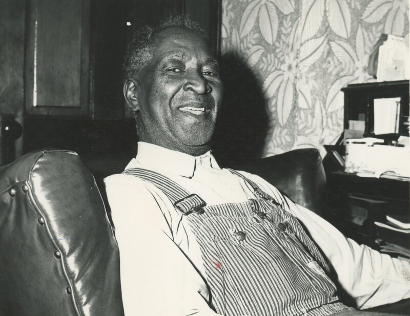 An older person sat in an arm chair smiling at the camera.