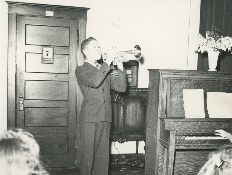 A young person stood next to a piano in a nice suit playing a trumpet in a room for people.