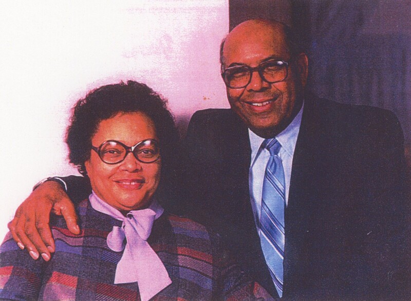 Two people sat next to eachother smiling for a photo while one has their arm wrapped around the other's shoulders.