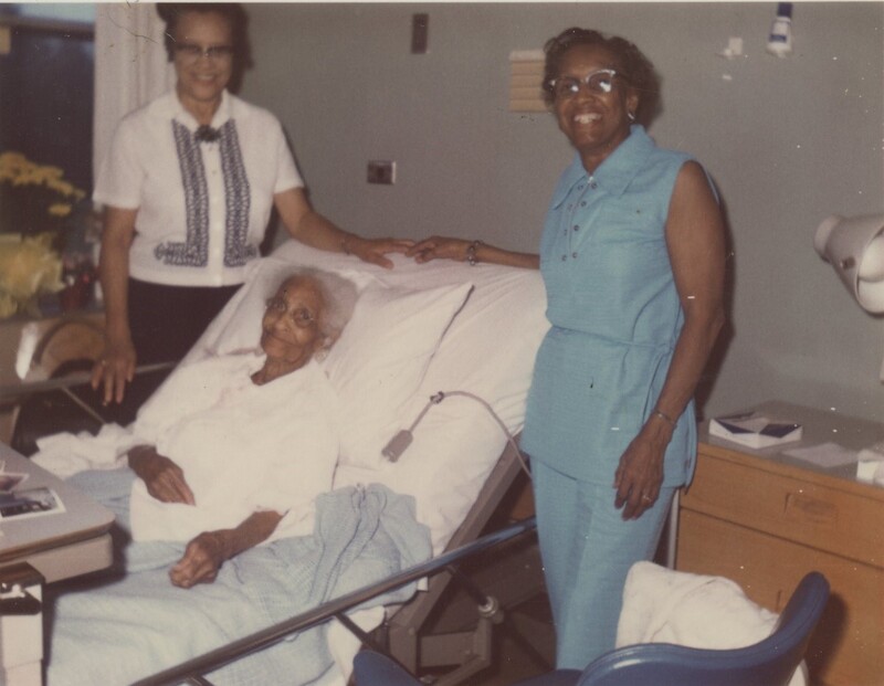 An older person laying down in a hospital bed with two other people stood around them all smiling for a photo.