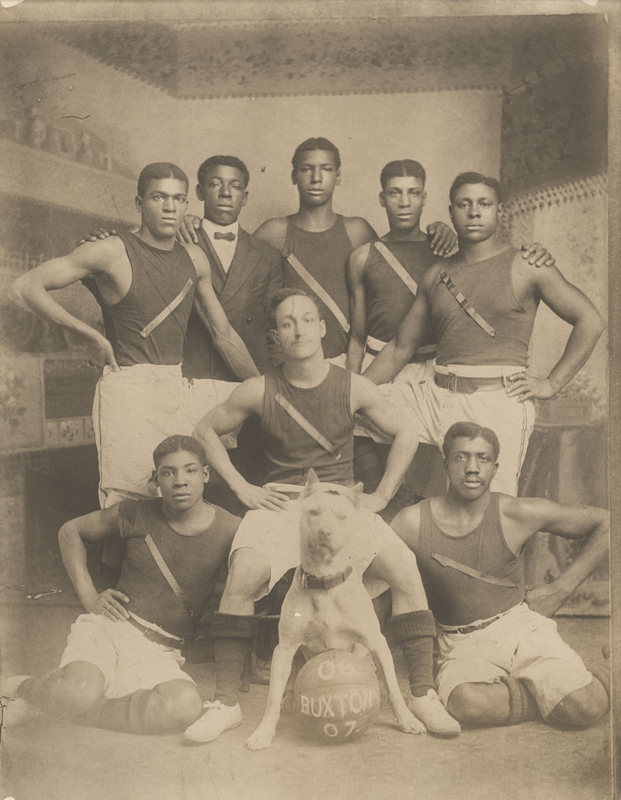 Photograph of members of the 8-member Buxton basketball team with dog as team mascot. Photograph is undated; however, the pictured basketball includes the text "06 - Buxton - 07" indicating the team played during the years 1906-1907.