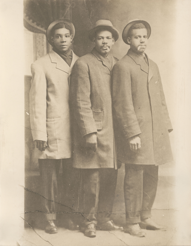 Photograph of 3 men wearing hats, overcoats, trousers, and shoes, taken inside a room. Photograph is undated.