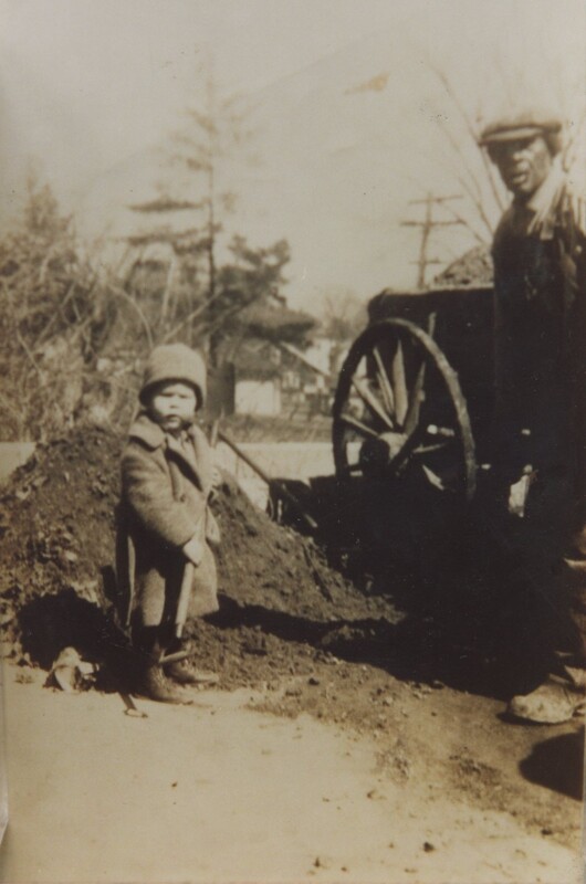 An older man and a little boy bundled up in a coat, scarf and hat stoodt infront of a pile of dirt and a carriage.