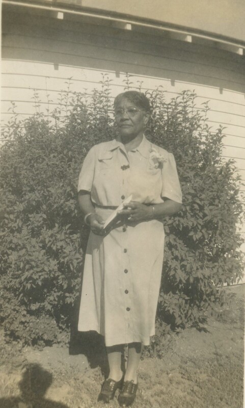An older person stood in front of a larger bush standing in a button down dress.