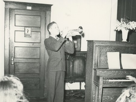 A young person stood next to a piano in a nice suit playing a trumpet in a room for people.