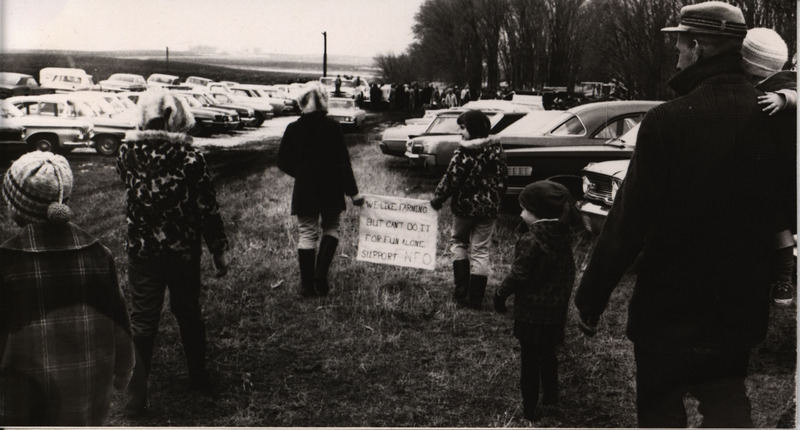 Photograph of a family walking through field with parked cars holding sign that reads, "We like farming but can't do it for fun alone. Support NFO."
