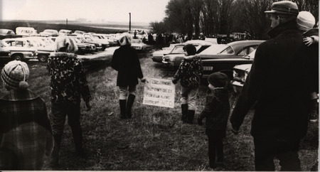 Photograph of a family walking through field with parked cars holding sign that reads, "We like farming but can't do it for fun alone. Support NFO."