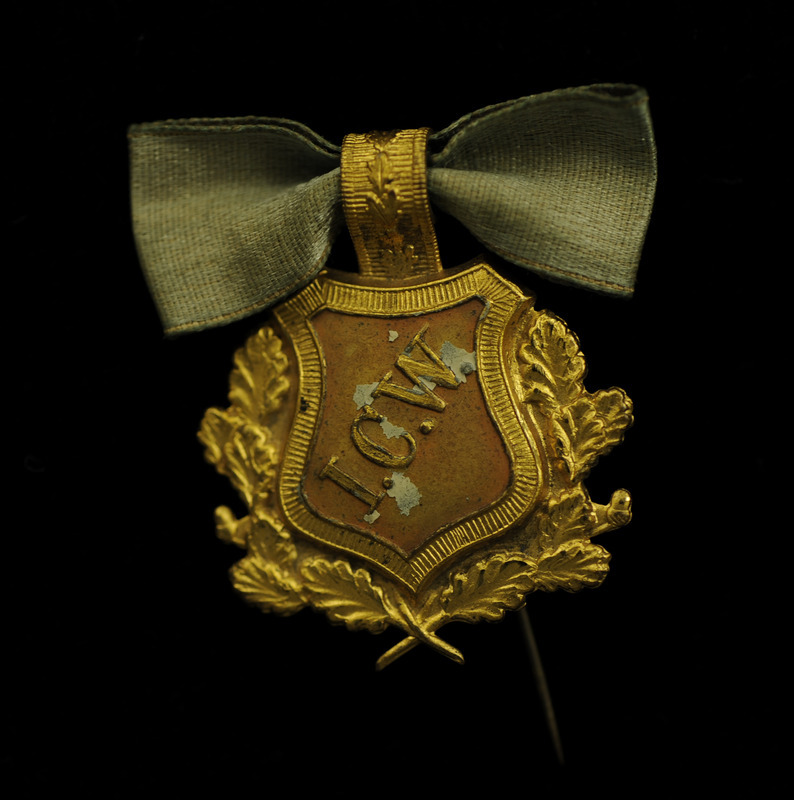 Decorative pin with a gold plated emblem that looks like a shield surrounded by an ornate leaf border. Front has raised lettering inscription of "ICW" in a diagonal. The top has a faded blue ribbon bow.