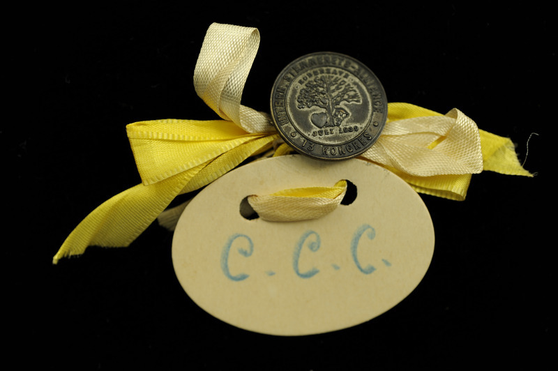 Silver, round button with "Intern. Stemmert Alliance 13 Kongress" around the edge "Kobenhaven", "July 1939," and a tree and a heart embossed on it. It is connected with white and yellow ribbons to an oval piece of white paper with "C.C.C." written in blue. The badge is from the Thirteenth Congress of the International Alliance of Women, Copenhagen, Denmark, July 8-14, 1939.
