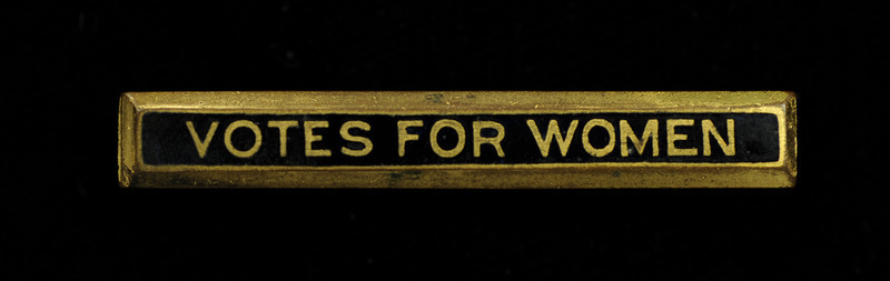 Gold, rectangular pin with "VOTES FOR WOMEN" inscribed in gold on a black inlay band on the front.