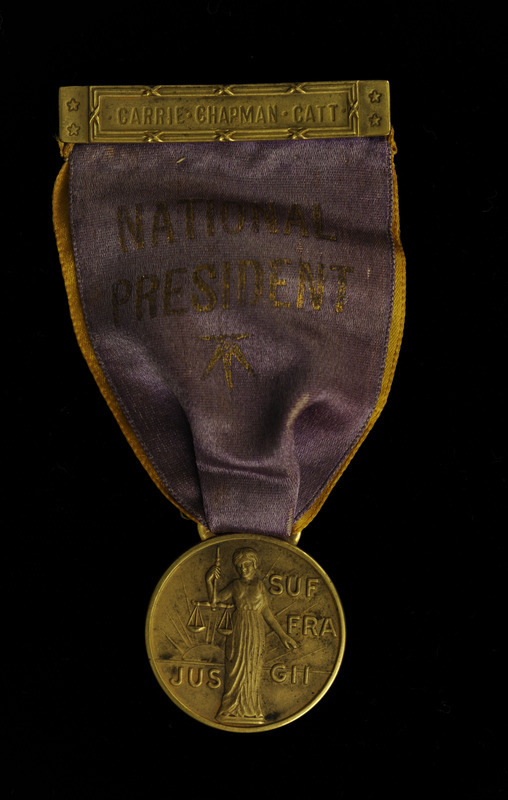 Ribbon consists of a small gold bar at top with "CARRIE CHAPMAN CATT" etched on the front and the back has the pin. Beneath the bar is a gold colored ribbon under a purple ribbon with "NATIONAL PRESIDENT" in gold lettering and a gold star underneath. Attached to the ribbons is a round, gold medal with an image of women holding scales in front of a rising sun and "Jus Suffragii" (The Right of Suffrage) in raised lettering.