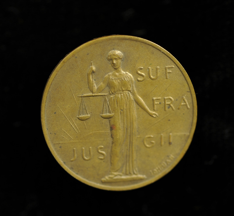 Round, gold medal with an image of women holding scales in front of a rising sun and "Jus Suffragii" (The Right of Suffrage) in raised lettering.