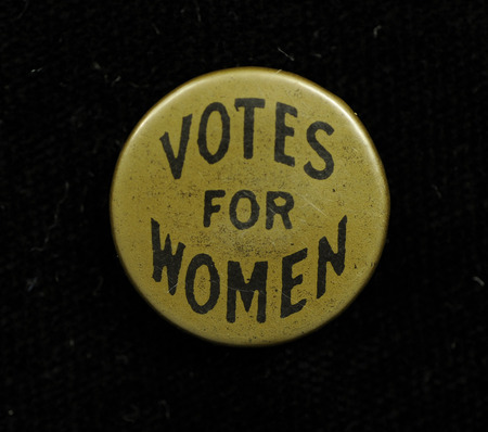 Round gold colored button with "VOTES FOR WOMEN" in black lettering on the front.