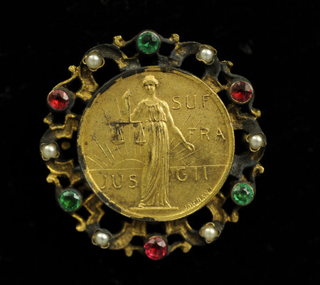 Round, gold pin with an image of women holding scales in front of a rising sun and "Jus Suffragii" (The Right of Suffrage) in raised lettering, surrounded by emeralds, pearls, and rubies.