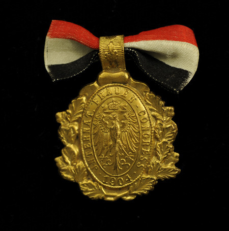 Gold, oval, badge surrounded by gold leaves with an image of an eagle and "Internat. Frauen Congress 1904" embossed on it; a red, white and blue bow is attached at the top. The badge is from the International Woman Suffrage Alliance Second Congress, which was held in Berlin, Germany, June 3-4, 1904.