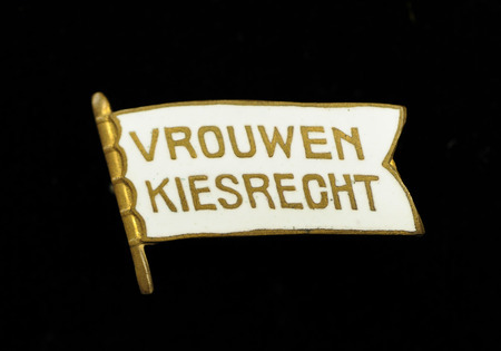 Small pin in the shape of a flag. Front has white background inscribed with "VROUWEN KIESRECHT" in gold. Vrouwenkiesrecht translates from Dutch to "Woman Suffrage" in English.