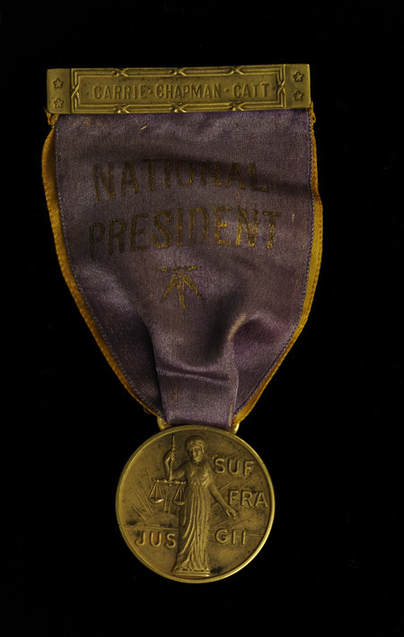 Ribbon consists of a small gold bar at top with "CARRIE CHAPMAN CATT" etched on the front and the back has the pin. Beneath the bar is a gold colored ribbon under a purple ribbon with "NATIONAL PRESIDENT" in gold lettering and a gold star underneath. Attached to the ribbons is a round, gold medal with an image of women holding scales in front of a rising sun and "Jus Suffragii" (The Right of Suffrage) in raised lettering.