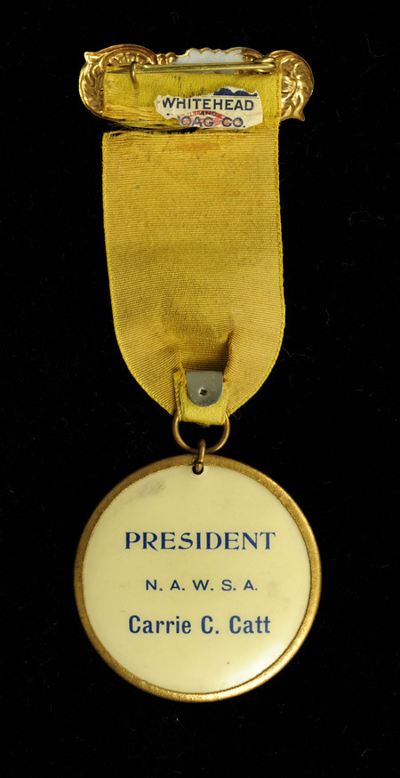 Ribbon includes a small gold decorative rectangle that is at the top. Attached is a yellow ribbon with "N. Y. W. S. A. 35th Annual Convention Hornellsville Oct. 20-23, 1903" in black letter. Attached at the bottom of the ribbon is a round photograph of Carrie Chapman Catt framed by a gold ring. The back of the image has "PRESIDENT N.A.W.S.A. Carrie C. Catt" in blue lettering.