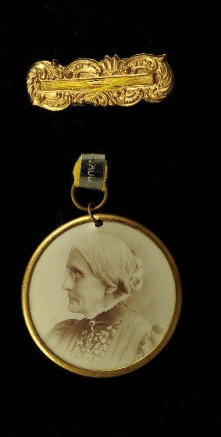 Incomplete badge with only the ornate gold rectangle that consists of the pin and remnants of a gold ribbon on it. There is round photograph of Susan B. Anthony framed by a gold ring. The back of the image has "Perfect equality of rights for women. Susan B. Anthony." inscribed on it.