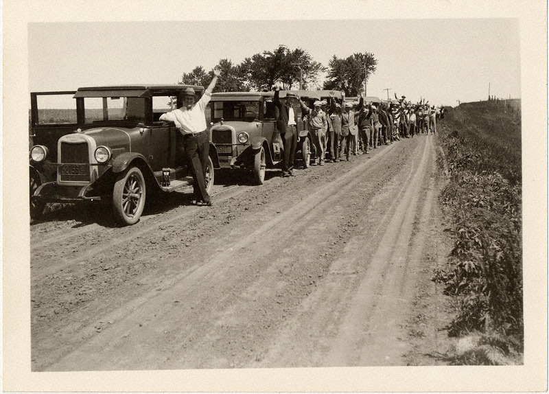 Extension Service with cars all in a line down a gravel road, the drivers waving.