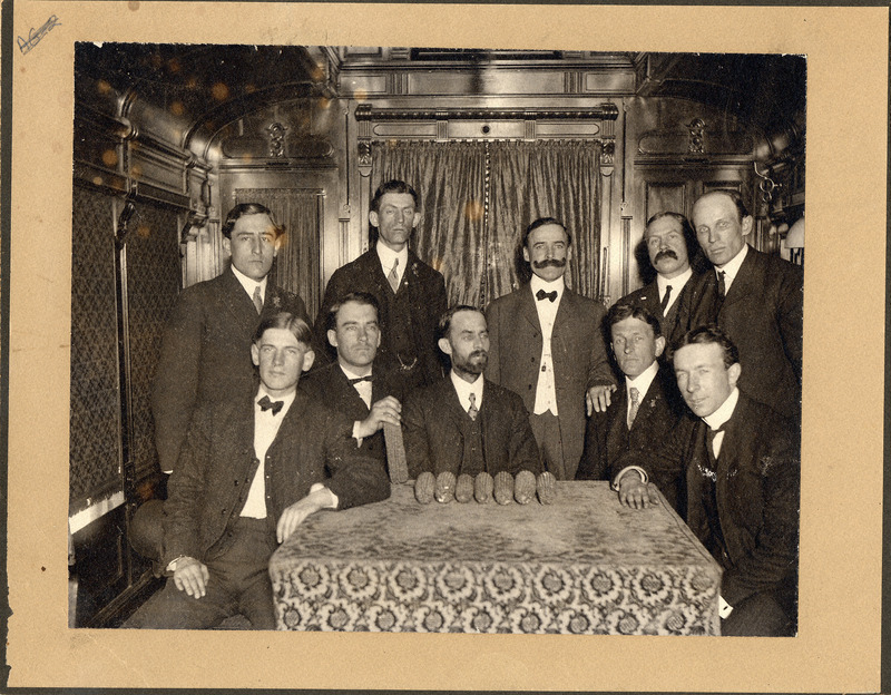 Photograph of Perry Holden in center with other men in corn train. Seven ear of corn lay before them on a table.
