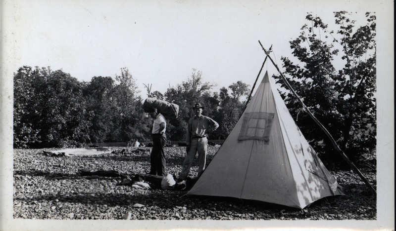 Frederic Leopold (in hat) and an unidentified man stand next to a tent. Behind them, a third person sits on the edge of a tree-lined stream.