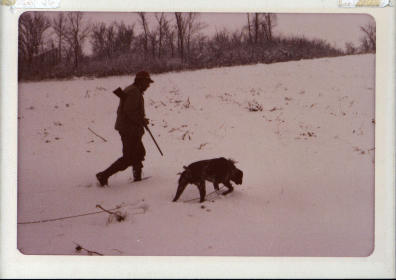 Frederic Leopold, carrying a rifle, walks across a snow covered field with a hunting dog, 1978.