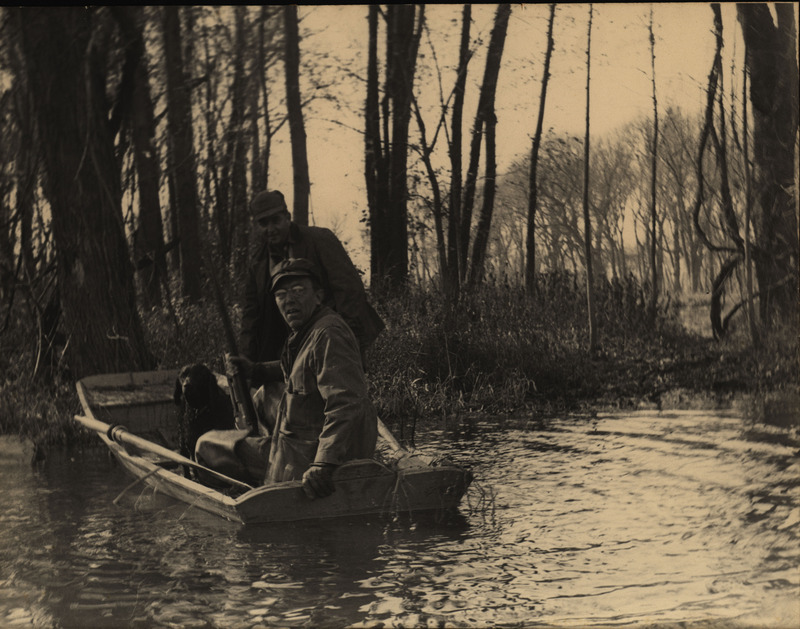 Frederic Leopold, seated and holding a rifle, and John Hale along with a hunting dog in a small paddle boat. The boat is situated near a tree-lined stretch of Two Key Island located nearby the Mississippi River above the Iowa River.